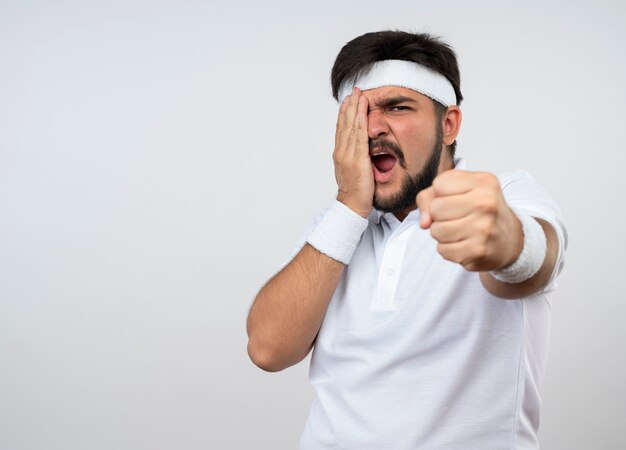 Jovem bravo e esportivo usando bandana e pulseira, segurando o punho e colocando a mão no rosto isolado na parede branca com espaço de cópia