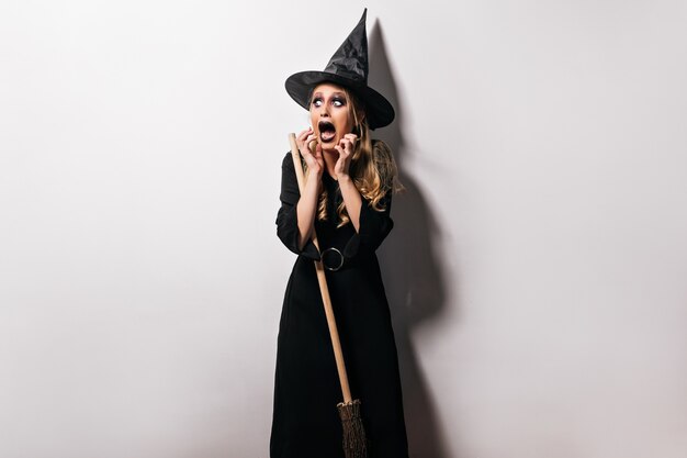 Jovem bruxa com chapéu, posando no halloween com expressão de rosto assustador. Foto interna da modelo feminina loira chocada com fantasia de assistente.