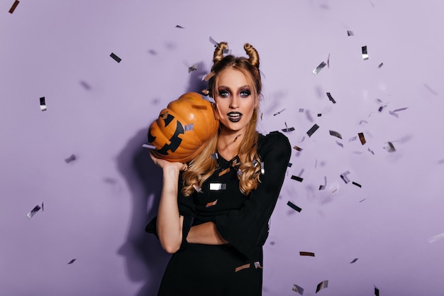 Foto grátis jovem bruxa confiante segurando abóbora de halloween. foto de menina bonita vampira em pé na parede roxa.