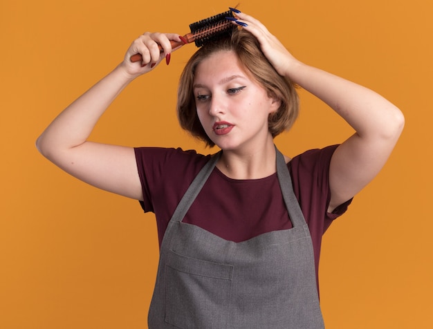 Jovem cabeleireira linda com avental penteando o cabelo com uma escova, olhando para o lado com uma cara séria em pé sobre uma parede laranja