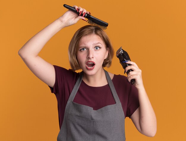 Jovem cabeleireira linda com avental segurando o aparador e uma escova de cabelo penteando o cabelo, parecendo confusa e surpresa em pé sobre uma parede laranja