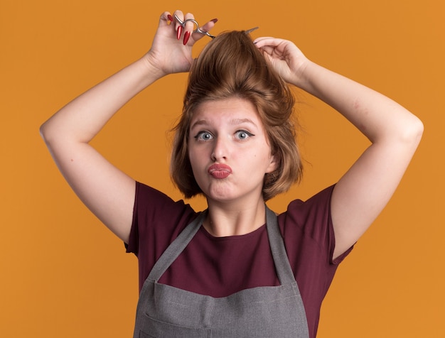 Foto grátis jovem cabeleireira linda com avental tentando cortar o cabelo com uma tesoura, parecendo confusa e surpresa em pé sobre uma parede laranja