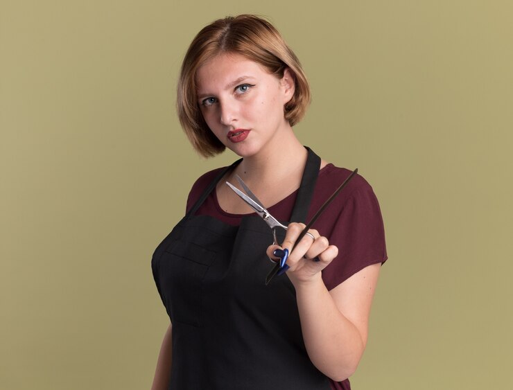 Foto grátis jovem cabeleireira linda de avental com tesoura e pente de cabelo olhando para frente com cara séria em pé sobre a parede verde