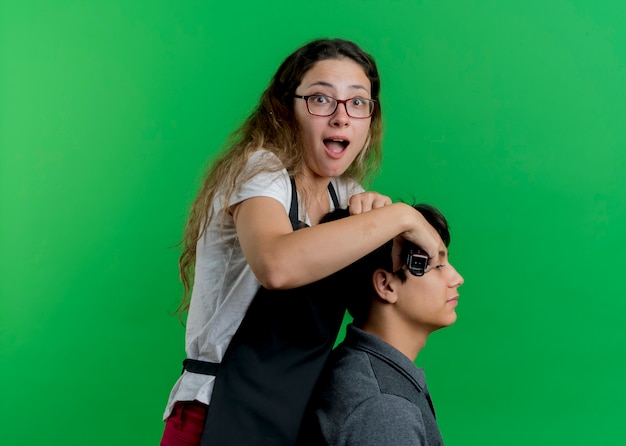 Foto grátis jovem cabeleireira profissional com avental cortando cabelo com aparador de cliente homem, olhando para frente sendo surpreendida em pé sobre a parede verde