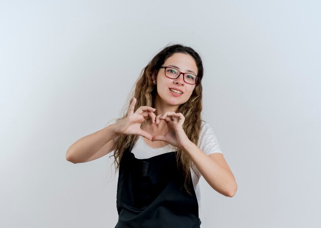 Jovem cabeleireira profissional com avental fazendo gestos de coração com os dedos sorrindo em pé sobre uma parede branca