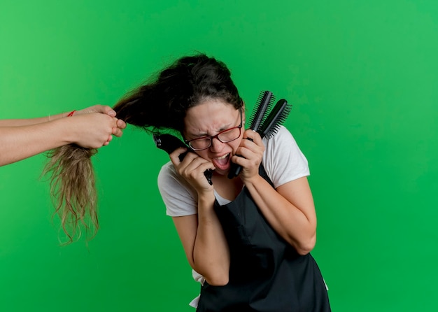 Foto grátis jovem cabeleireira profissional com avental segurando escovas de cabelo e sentindo-se penteada porque alguém puxando seu cabelo