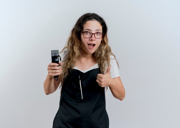 Jovem cabeleireira profissional com avental segurando o aparador, olhando para a frente, sorrindo e mostrando os polegares em pé sobre uma parede branca