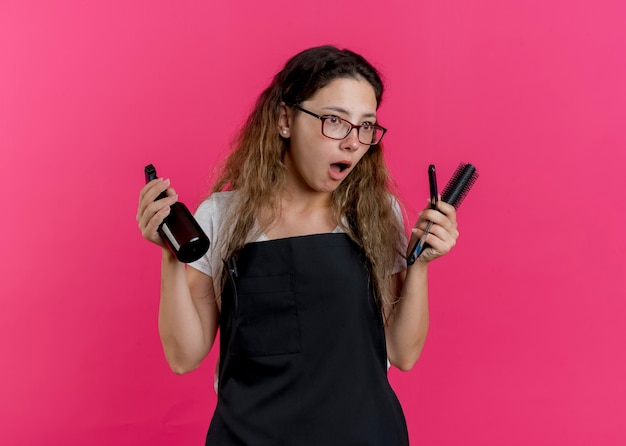 Foto grátis jovem cabeleireira profissional com avental segurando spray, escova de cabelo e lâmina de barbear, parecendo de lado surpresa e surpresa