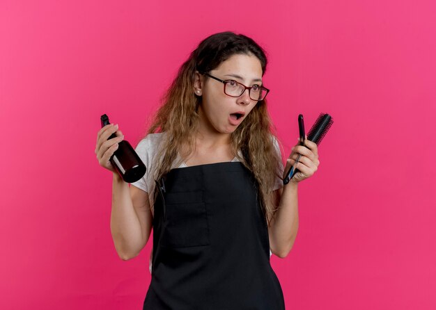 Jovem cabeleireira profissional com avental segurando spray, escova de cabelo e lâmina de barbear, parecendo de lado surpresa e surpresa