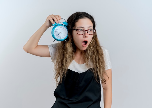 Foto grátis jovem cabeleireira profissional com avental segurando um despertador, olhando de lado e se surpreendendo em pé sobre uma parede branca