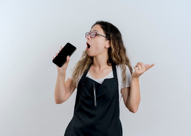 Jovem cabeleireira profissional com avental segurando um smartphone e usando-o como microfone cantando se divertindo em pé sobre uma parede branca