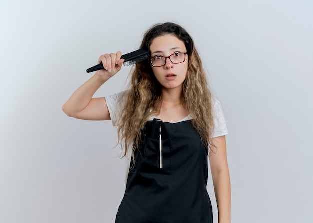 Jovem cabeleireira profissional com avental segurando uma escova de cabelo, penteando o cabelo, olhando para a frente, confusa em pé sobre uma parede branca