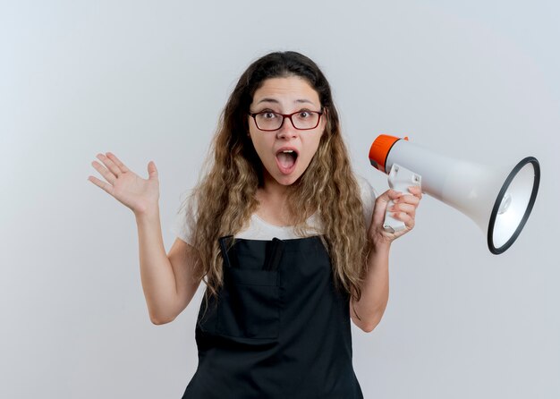 Jovem cabeleireira profissional de avental segurando um megafone, olhando para a frente, gritando e sendo confusa em pé sobre uma parede branca