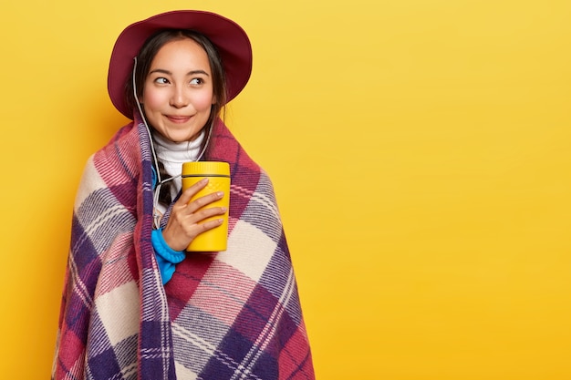 Foto grátis jovem, calma e agradável, garota asiática em pé envolta em uma manta quentinha, segurando uma xícara de café para viagem, usa chapéu, aproveita o tempo para descansar