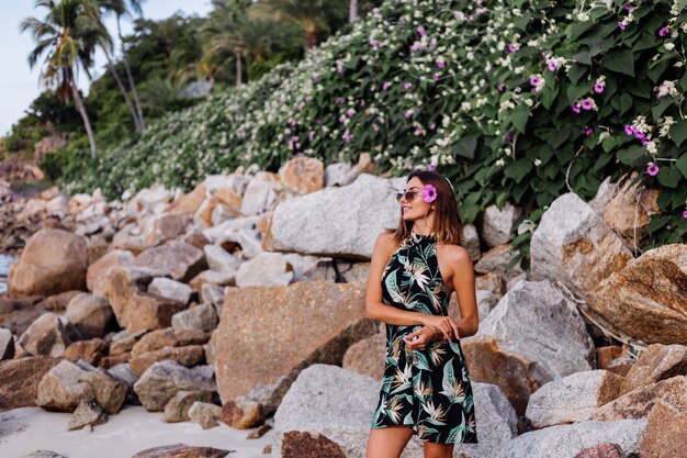 Jovem calma e tatuada em um vestido curto de estampa tropical de verão em uma praia rochosa com arbusto verde e flores rosa roxas