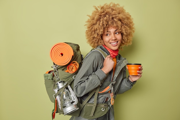 Jovem campista feminina positiva bebe água sente sede depois de andar muito tempo carrega mochila com lanterna karemat enrolada e outros equipamentos isolados sobre fundo verde Estilo de vida de aventura