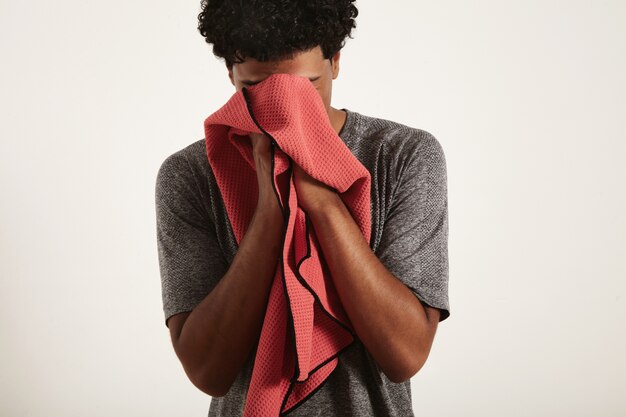 Jovem cansado e desapontado em forma de atleta negro em camisa cinza, enxugando o suor do rosto com uma toalha waffle vermelha sobre branco