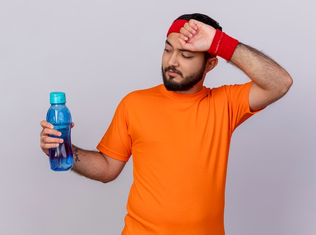 Jovem cansado e esportivo usando bandana e pulseira, segurando uma garrafa de água e enxugando a testa com a mão isolada no fundo branco