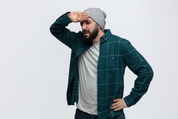 Jovem cansado usando chapéu de inverno olhando para o lado mantendo a mão na cintura enquanto mantém a outra mão na testa isolada no fundo branco