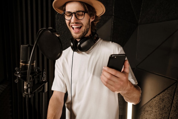 Jovem cantor masculino atraente com smartphone cantando emocionalmente no estúdio de gravação moderno