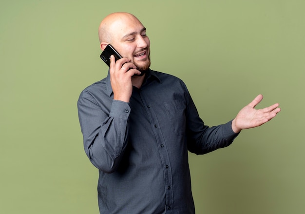 Jovem careca alegre homem de call center olhando para o lado falando no telefone e mostrando a mão vazia isolada em verde oliva