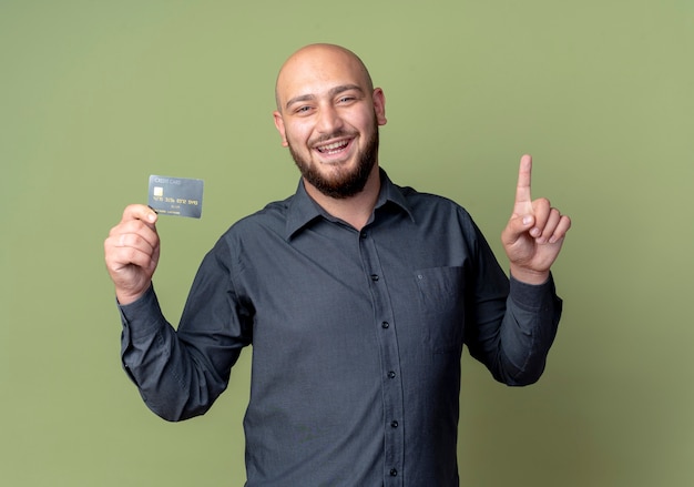 Foto grátis jovem careca alegre homem de call center segurando um cartão de crédito e levantando o dedo isolado em verde oliva