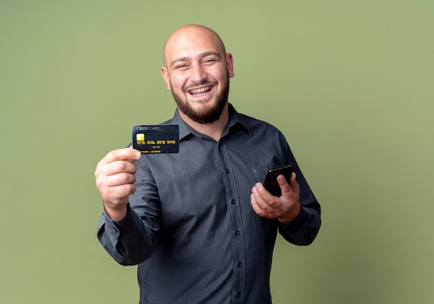 Jovem careca alegre homem de call center segurando um telefone celular e estendendo o cartão de crédito isolado em verde oliva
