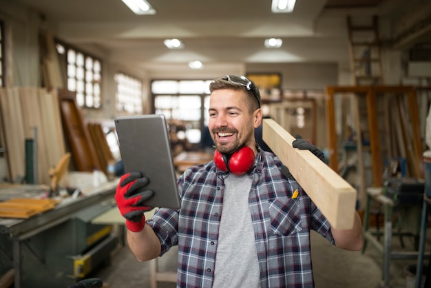 Jovem carpinteiro profissional segurando material de madeira e olhando para um tablet na oficina de carpintaria
