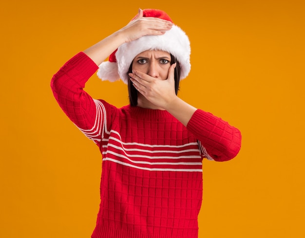 Jovem carrancuda com chapéu de Papai Noel, olhando para a câmera, segurando a cabeça, cobrindo a boca com a mão isolada em fundo laranja