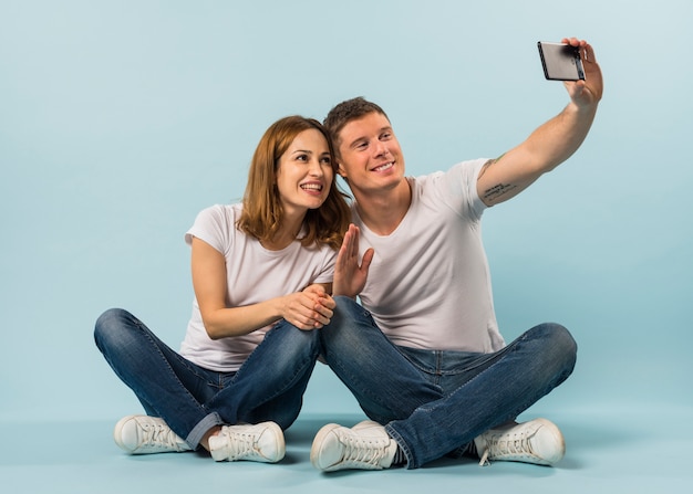 Jovem casal acenando a mão dela tomando selfie no smartphone contra o pano de fundo azul