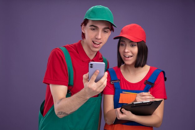 Jovem casal alegre com uniforme de trabalhador da construção civil e boné segurando a garota do celular segurando um lápis e uma prancheta, ambos olhando para o celular isolado na parede roxa