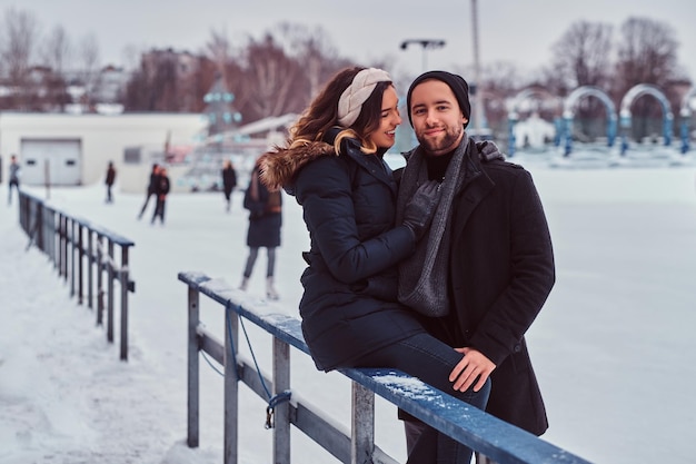Jovem casal apaixonado, namora na pista de gelo, uma garota sentada em um guardrail e abraçando o namorado.