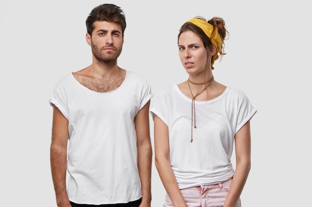 Jovem casal apaixonado tem expressões faciais desagradáveis, olha com aversão, insatisfeito com os maus resultados de seu trabalho, usa camiseta branca, bandana amarela