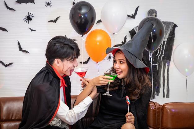 Jovem casal asiático feliz vestindo fantasia de halloween vampiro e bruxa comemorando em festa segurando e tilintando copo de vinho olhando para a câmera