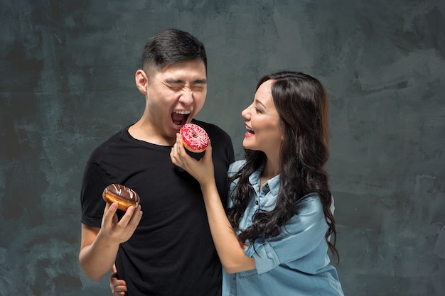 Jovem casal asiático gosta de comer rosquinha doce e colorida em estúdio cinza