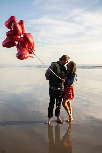 Jovem casal beijando na praia ao pôr do sol enquanto segura balões em forma de coração