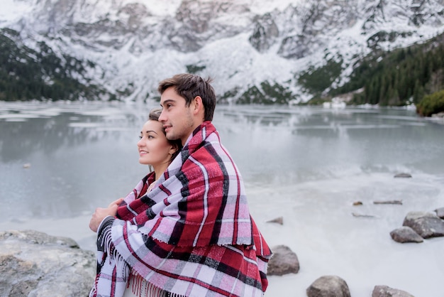 Jovem casal coberto com cobertor brilhante está de pé na frente do lago das montanhas congelado