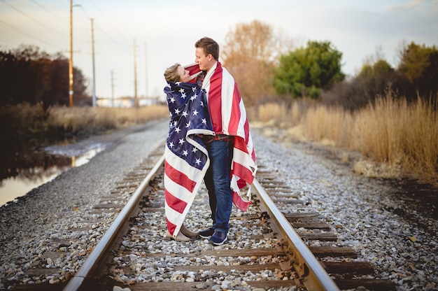 Jovem casal com a bandeira americana pendurada nos ombros em pé na ferrovia