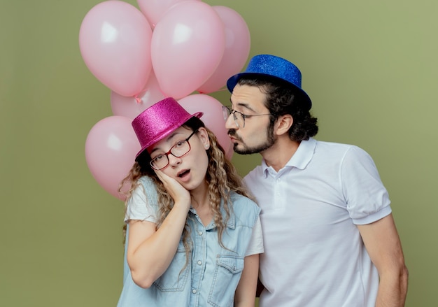 Jovem casal com chapéu rosa e azul parado na frente da garota dos balões colocando a mão na bochecha
