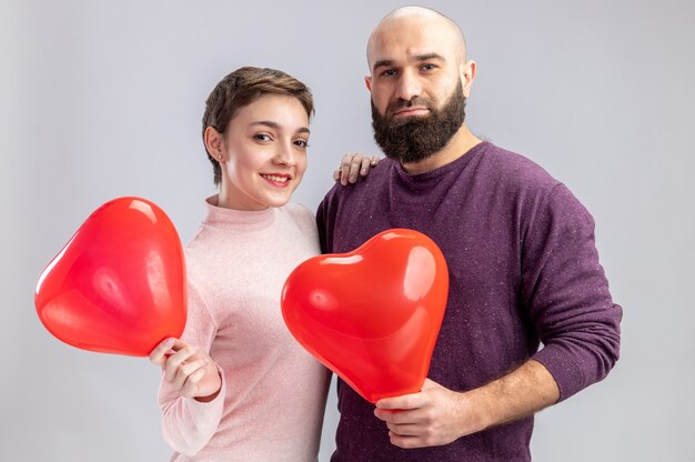 jovem casal com roupas casuais, homem e mulher segurando balões em forma de coração, olhando para a câmera, feliz e alegre, sorrindo celebrando o dia dos namorados em pé sobre uma parede branca