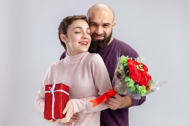 jovem casal com roupas casuais mulher com cabelo curto com presente e homem barbudo com buquê de rosas vermelhas abraçando feliz no amor comemorando o dia dos namorados em pé sobre um fundo branco