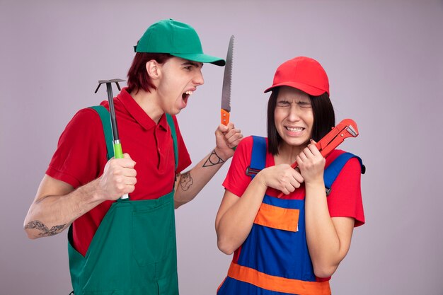 Jovem casal com uniforme de trabalhador da construção civil e boné menina assustada segurando a chave de cachimbo com os olhos fechados cara bravo segurando enxada e serra olhando para a menina gritando