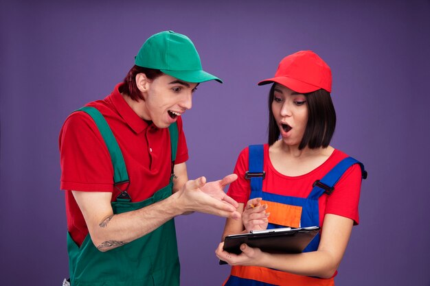 Jovem casal com uniforme de trabalhador da construção civil e boné surpreendeu a garota segurando um lápis e uma prancheta, cara animado apontando com as mãos para a prancheta, ambos olhando para a prancheta