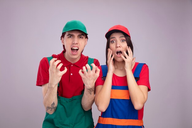 Jovem casal com uniforme de trabalhador da construção civil e boné zangado, mostrando as mãos vazias, garota preocupada, mantendo as mãos perto do rosto, ambos olhando para a câmera isolada na parede branca