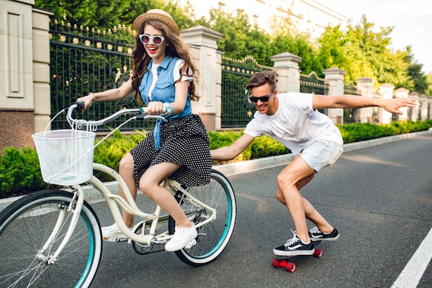 Foto grátis jovem casal de adolescentes bonitos se divertindo no verão, no pôr do sol na estrada. menina bonita com cabelo longo cacheado no chapéu, dirigindo uma bicicleta, cara bonito mantém a bicicleta e anda de skate.