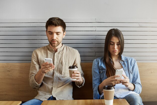 Jovem casal de estudantes sentados em um encontro em fast-food, tomando café, olhando em telefones celulares, ignorando-se após discussão