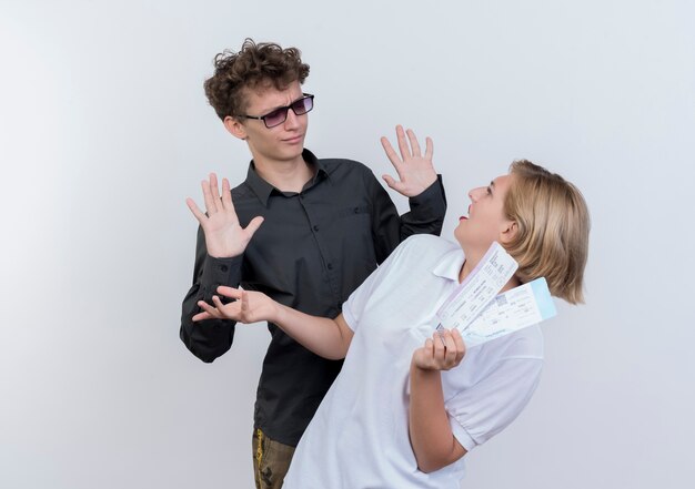 Jovem casal de turistas confuso e descontente, olhando para a namorada com passagens aéreas em mãos em pé sobre uma parede branca