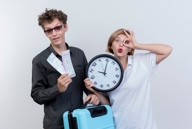 Jovem casal de turistas feliz segurando as passagens aéreas e a mala com a mulher segurando o relógio de parede fazendo o bem.