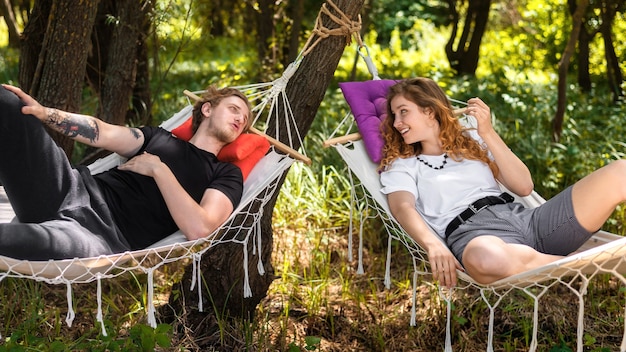Jovem casal deitado em redes, olhando um para o outro e sorrindo. vegetação ao redor. glamping
