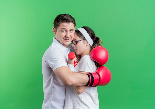 Jovem casal desportivo com luvas de boxe abraçando a namorada feliz e positivo em pé sobre a parede verde